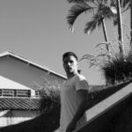 Black and white portrait of a man looking contemplatively, set against palm trees and a house background.